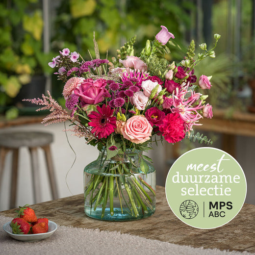 Duurzaam roze boeket met rozen, gerbera’s en lisianthus in fuchsia en zachtroze tinten in glazen vaas in een groene omgeving