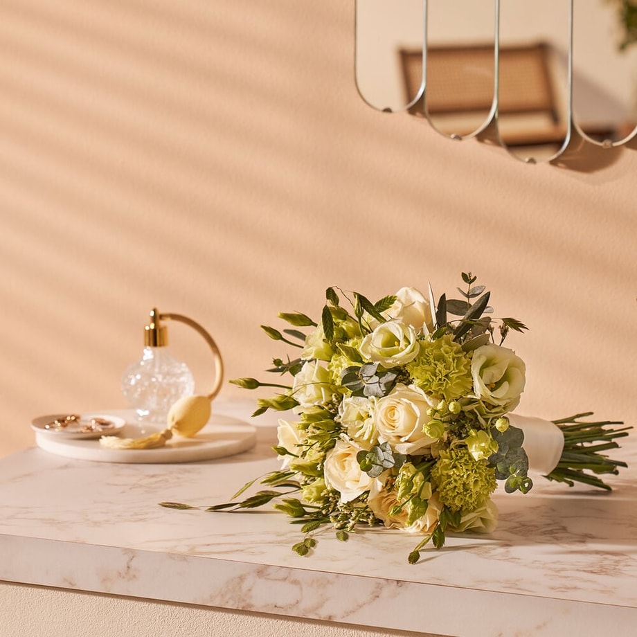 Elegant bruidsboeket met witte rozen en groene accenten, liggend op een marmeren tafel in een lichte, stijlvolle kamer.