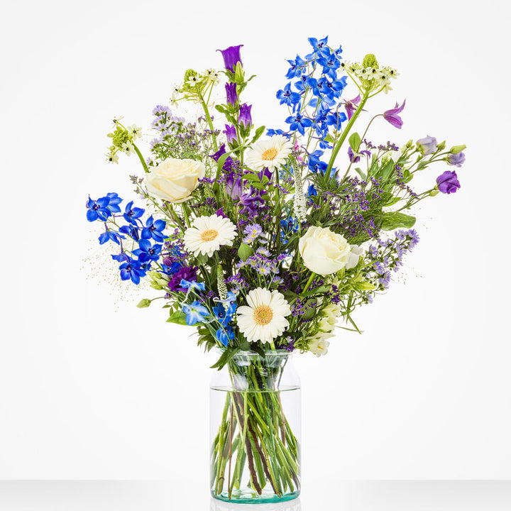 Groot boeket in een glazen vaas met witte rozen, blauwe bloemen en paarse accenten.