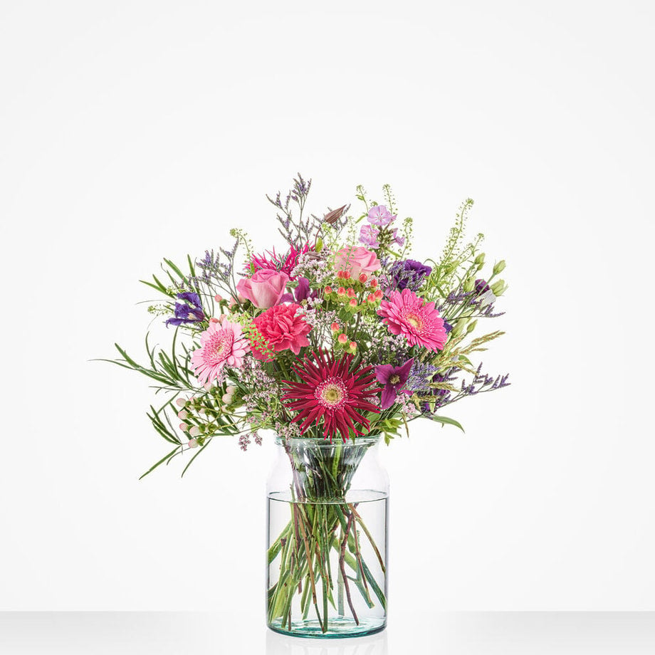 Middelgroot verjaardagboeket in een glazen vaas met roze, paarse en rode bloemen tegen een witte achtergrond.