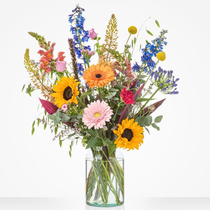 Groot zomerboeket in een glazen vaas met zonnebloemen, gerbera's, roze anjers, blauwe ridderspoor en paarse agapanthus tegen een witte achtergrond.