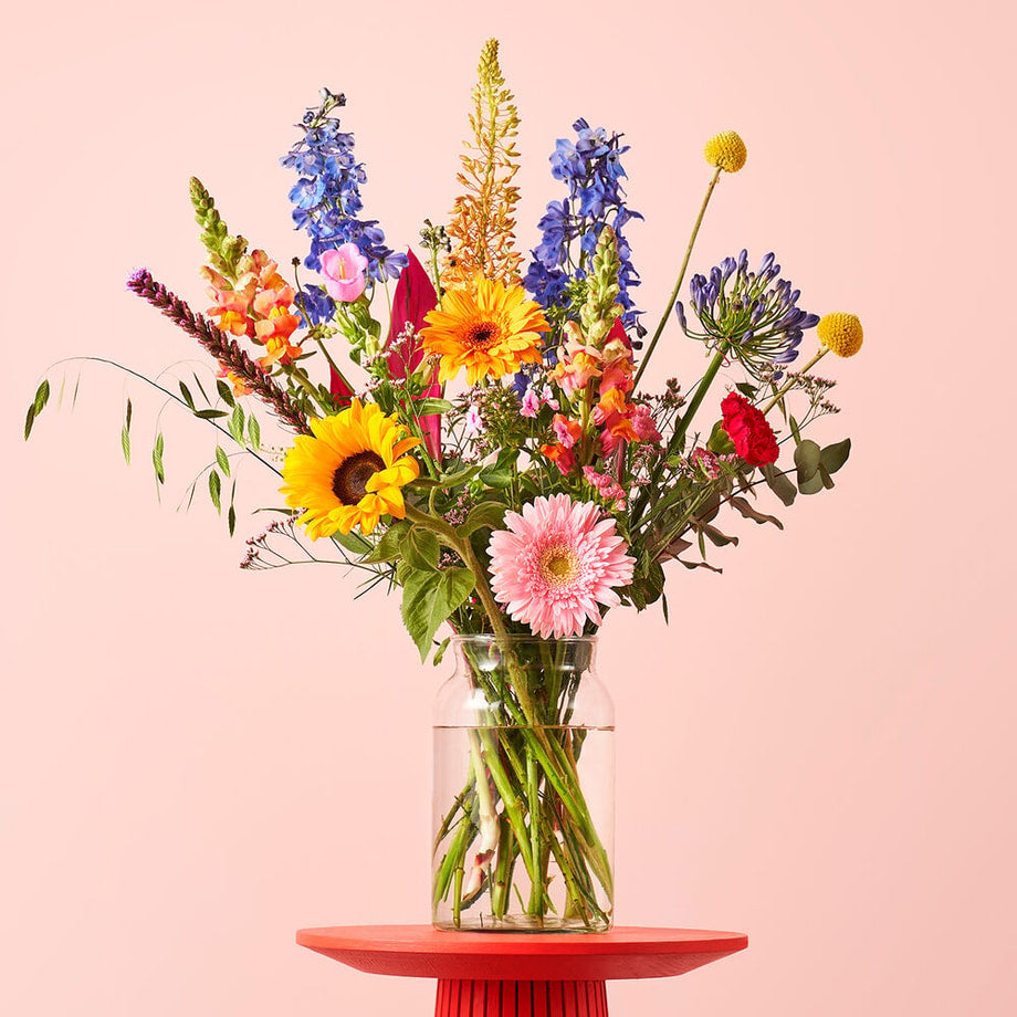 Zomerboeket in een vaas op een rode tafel tegen een roze achtergrond, met zonnebloemen, gerbera's, roze anjers, blauwe ridderspoor en paarse agapanthus.