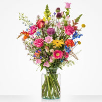 Groot fleurig boeket in een glazen vaas met roze rozen, gerbera's, gele bloemen, paarse allium, blauwe ridderspoor en oranje bloemen tegen een witte achtergrond.
