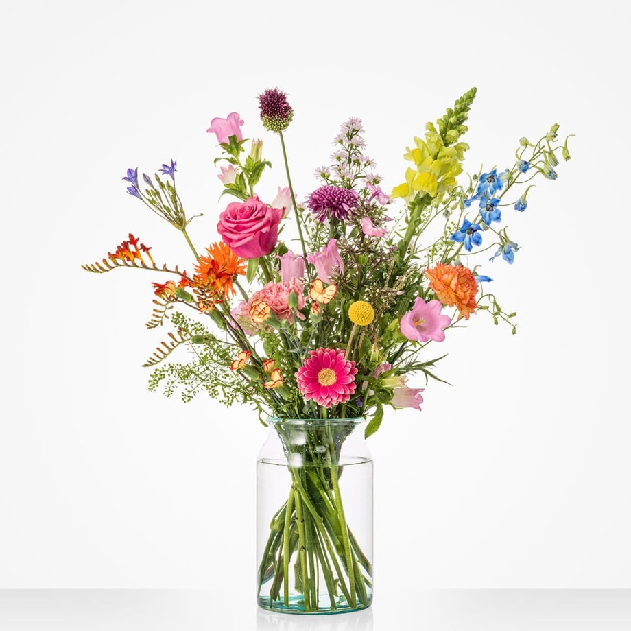 Klein fleurig boeket in een glazen vaas met roze rozen, gerbera's, gele bloemen, paarse allium, blauwe ridderspoor en oranje bloemen tegen een witte achtergrond.