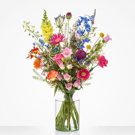 Middelgroot fleurig boeket in een glazen vaas met roze rozen, gerbera's, gele bloemen, paarse allium, blauwe ridderspoor en oranje bloemen tegen een witte achtergrond.