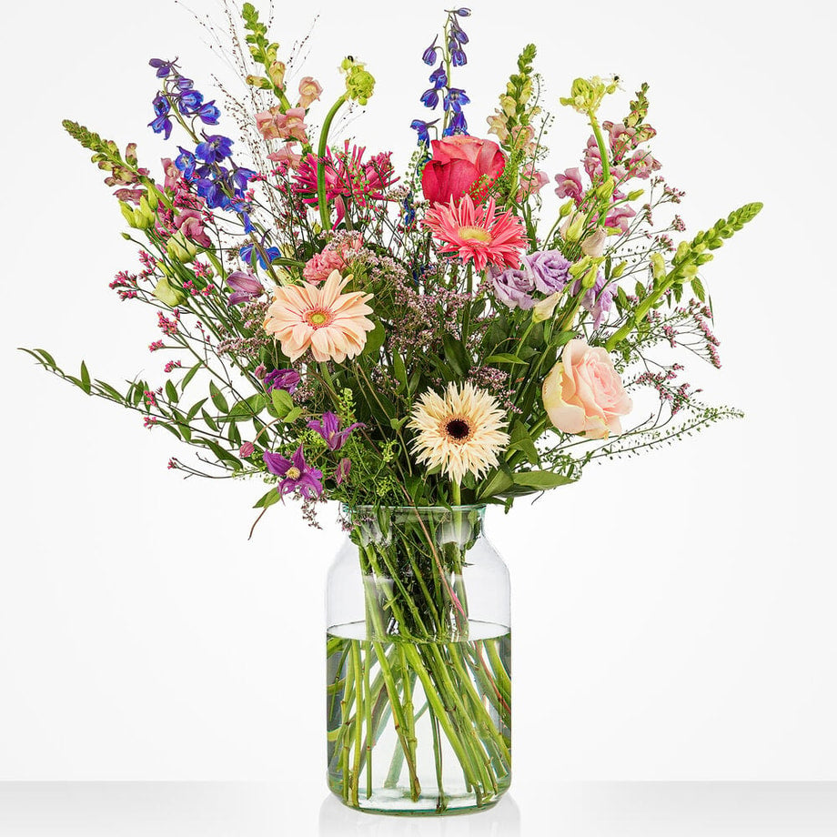 Groot pastelboeket in een glazen vaas met roze rozen, gerbera's, blauwe ridderspoor, lila bloemen en witte bloemen tegen een witte achtergrond.