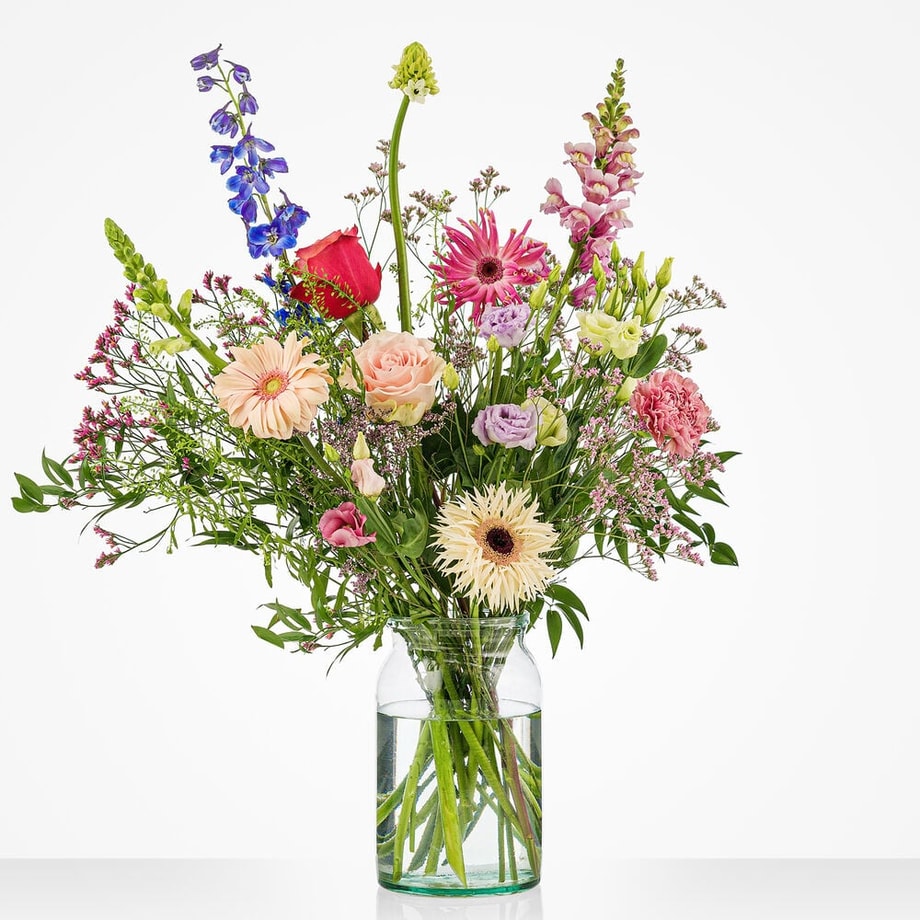 Klein pastelboeket in een glazen vaas met roze rozen, gerbera's, blauwe ridderspoor, lila bloemen en witte bloemen tegen een witte achtergrond.