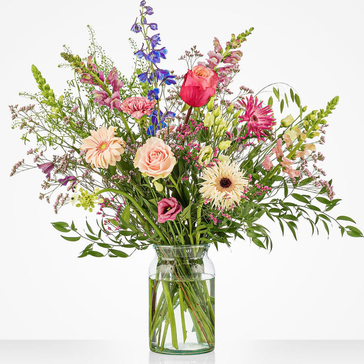 Middelgroot pastelboeket in een glazen vaas met roze rozen, gerbera's, blauwe ridderspoor, lila bloemen en witte bloemen tegen een witte achtergrond.