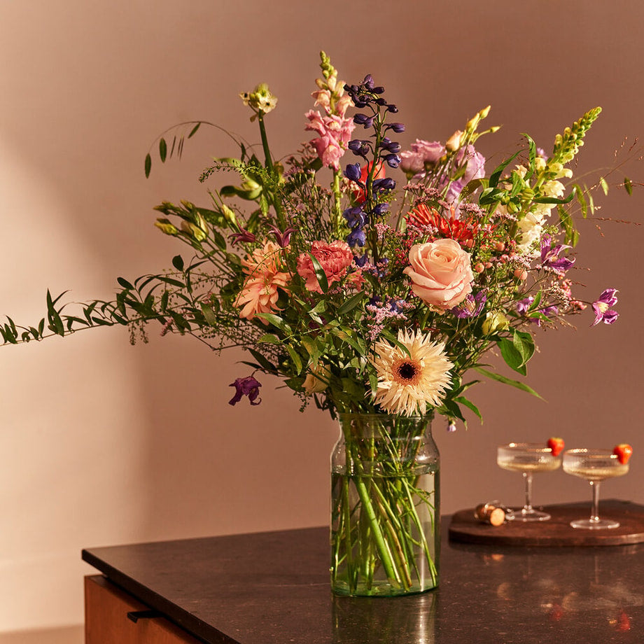 Pastelboeket in een vaas op een donkere tafel in een interieursetting, met roze rozen, gerbera's, blauwe ridderspoor, lila bloemen en witte bloemen.