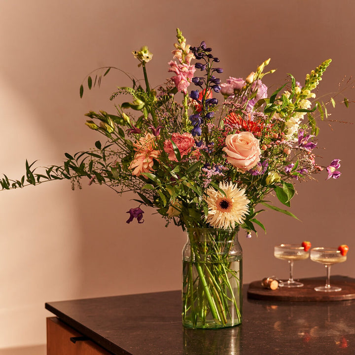 Pastelboeket in een vaas op een donkere tafel in een interieursetting, met roze rozen, gerbera's, blauwe ridderspoor, lila bloemen en witte bloemen.