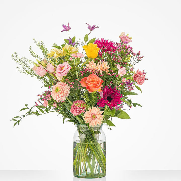 Groot boeket met een mix van kleurrijke bloemen, zoals felroze gerbera’s, oranje rozen en roze anjers, in een hoge glazen vaas.