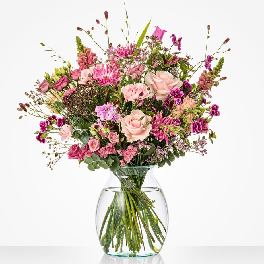 Groot boeket voor een trouwdag met roze rozen, paarse bloemen en diverse groene takken in een glazen vaas.