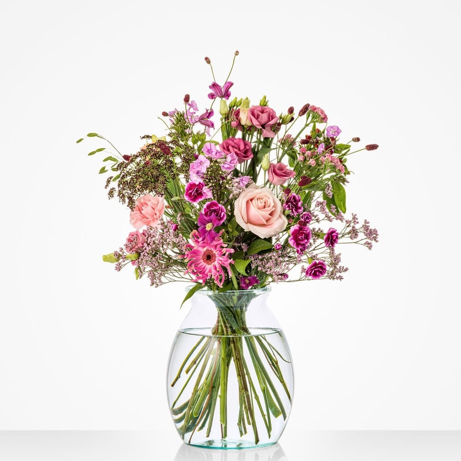 Klein boeket voor een trouwdag met zachte roze rozen, paarse bloemen en groen blad in een ronde glazen vaas.