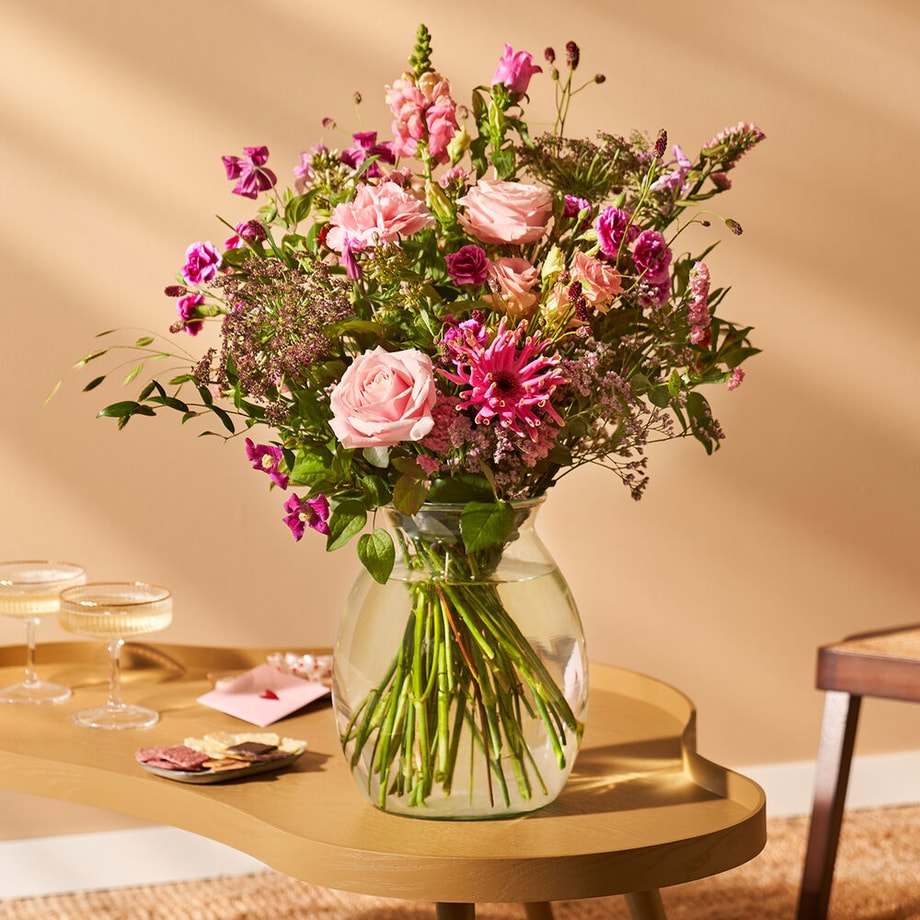 Sfeervolle setting met een groot boeket van roze en paarse bloemen in een glazen vaas op een bijzettafel, omringd door glazen en een kaart.