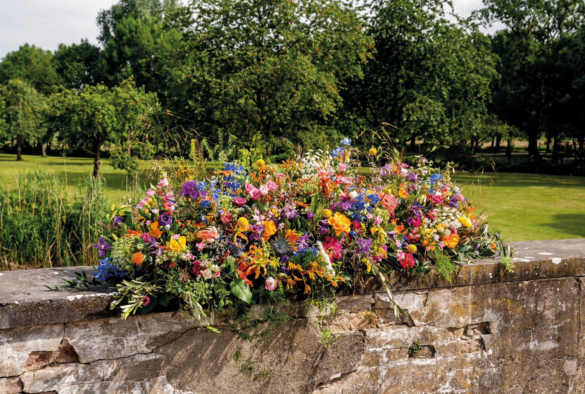 Gekleurd kistbedekking rouwstuk met diverse bloemen zoals gerbera’s, rozen en delphinium op stenen muur in groene omgeving.