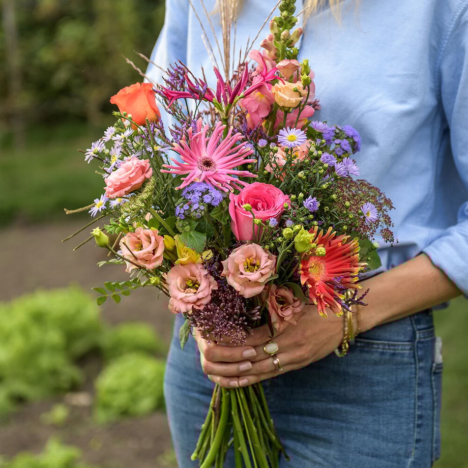 Vrouw in blauwe blouse houdt een kleurrijk boeket met rozen, gerbera’s en veldbloemen in haar handen, in een groene, natuurlijke omgeving.