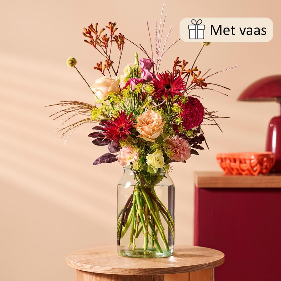 Herfstboeket met vaas, bestaande uit roze rozen, rode chrysanten en seizoensgroen, sfeervol gestyled op een houten tafeltje.