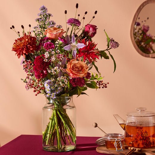 Herfstboeket met rozen, rode dahlia’s en paarse bloemen in glazen vaas, sfeervol gestyled.