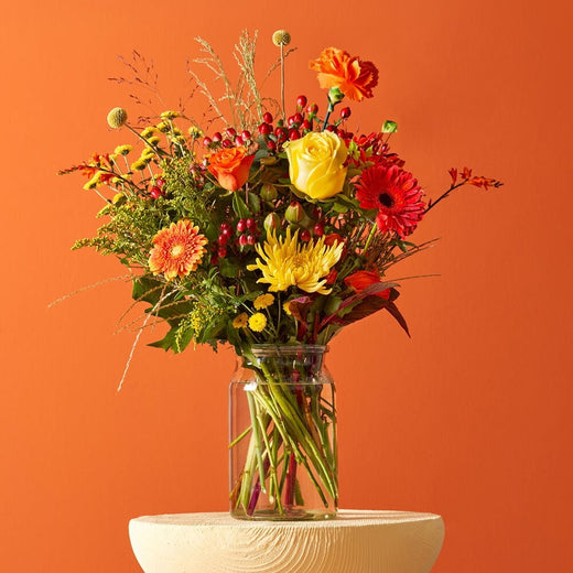 Herfstboeket met gele rozen, oranje gerbera’s en rode bessen in een glazen vaas tegen een oranje achtergrond.