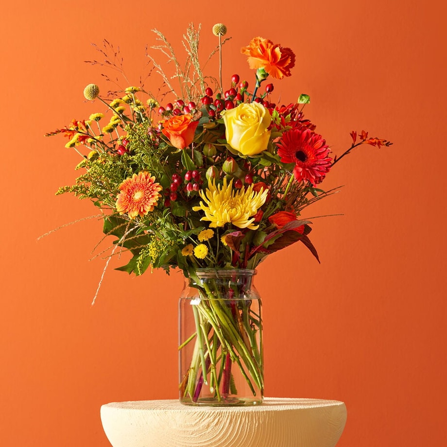 Herfstboeket met gele rozen, oranje gerbera’s en rode bessen in een glazen vaas tegen een oranje achtergrond.