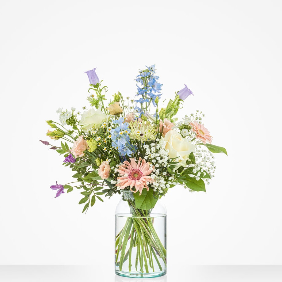 Middelgroot boeket in zachte tinten met bloemen zoals roze gerbera’s, blauwe ridderspoor en witte rozen in een glazen vaas.