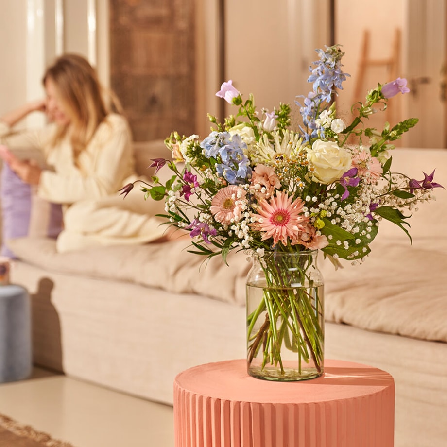 Sfeervolle setting met een zacht gekleurd boeket van rozen, gerbera’s, en veldbloemen in een glazen vaas op een roze tafel, met een vrouw op de achtergrond zittend op een bank.