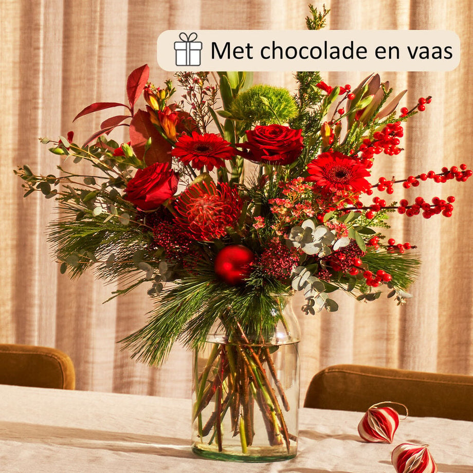 Kerstboeket met rode rozen, gerbera’s, bessen en dennengroen in glazen vaas, inclusief chocoladecadeau.
