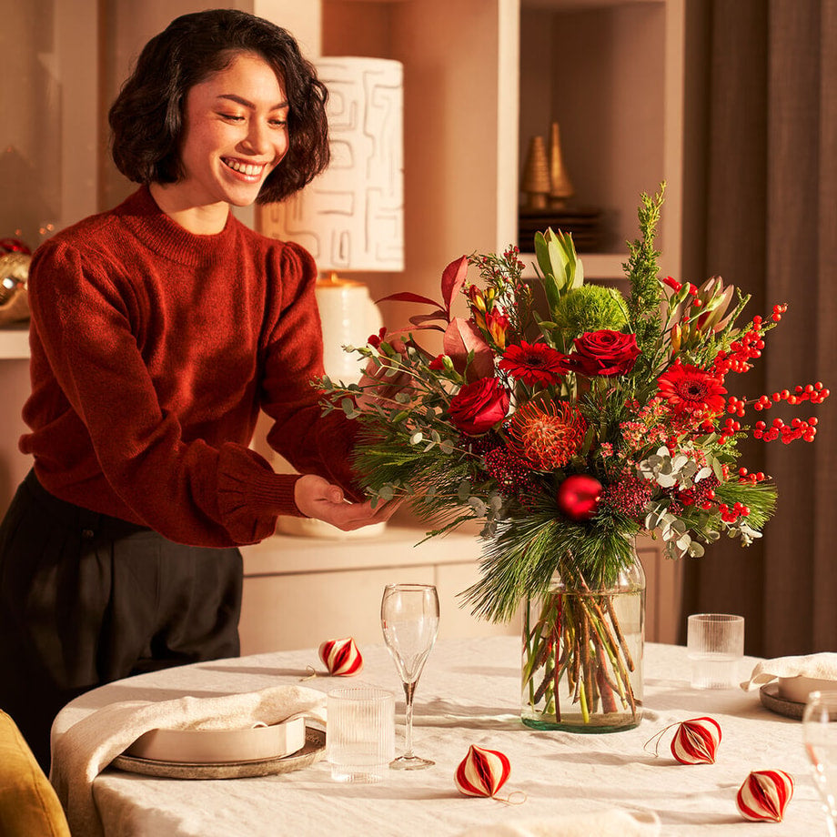 Vrouw plaatst een kerstboeket met rode rozen, gerbera’s en dennengroen op een feestelijk gedekte tafel.