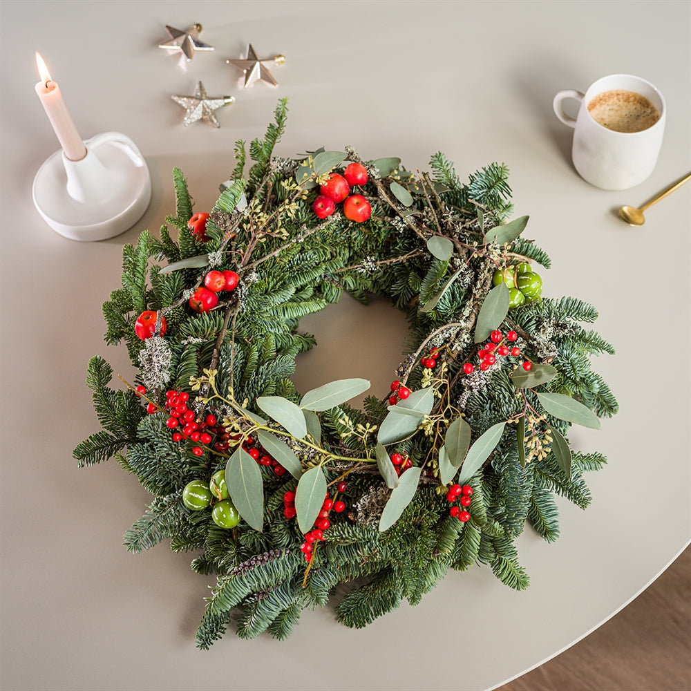 Kerstkrans met rode bessen, eucalyptus en dennengroen, gestyled met kaars en sterdecoraties op een tafel.