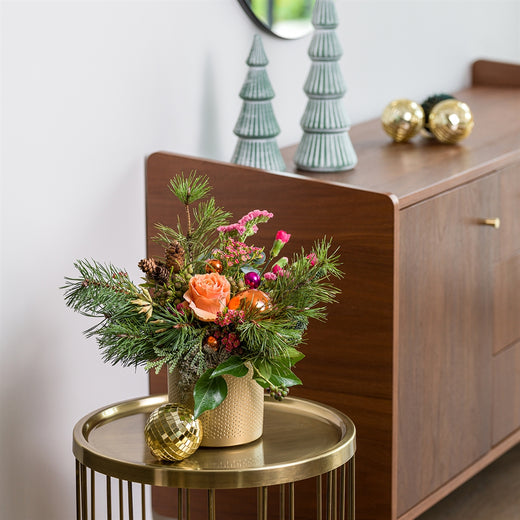 Kerststuk met oranje roos, dennengroen en kerstdecoraties in een gouden pot, gestyled op een ronde bijzettafel.