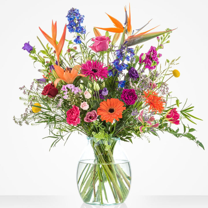 Middelgroot boeket met een levendige mix van bloemen zoals Strelitzia’s, gerbera’s, rozen en ridderspoor in een ronde glazen vaas.