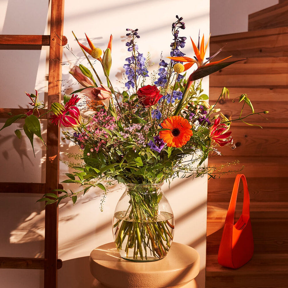 Sfeervolle hoek met een kleurrijk boeket van exotische bloemen zoals Strelitzia’s en gerbera’s in een glazen vaas, verlicht door zonnestralen in een interieursetting.