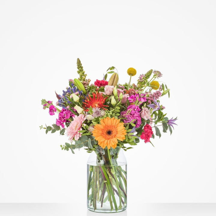 Middelgroot kleurrijk boeket met bloemen zoals oranje gerbera’s, roze rozen, paarse ridderspoor en andere felle bloemen in een glazen vaas.