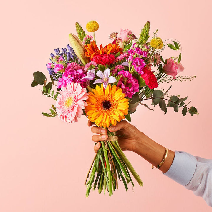 Hand die een kleurrijk boeket vasthoudt met bloemen zoals een gele gerbera, roze rozen, paarse bloemen en andere kleurrijke bloemen, tegen een zachte roze achtergrond.