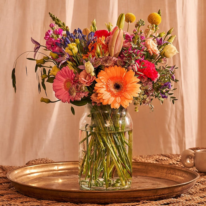 Gezellige setting met een kleurrijk boeket van bloemen zoals gerbera’s, rozen en lelies in een glazen vaas, geplaatst op een houten dienblad.