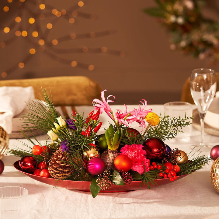 Kleurrijk kerststuk met bloemen, kerstballen en dennengroen op een feestelijk gedekte tafel.