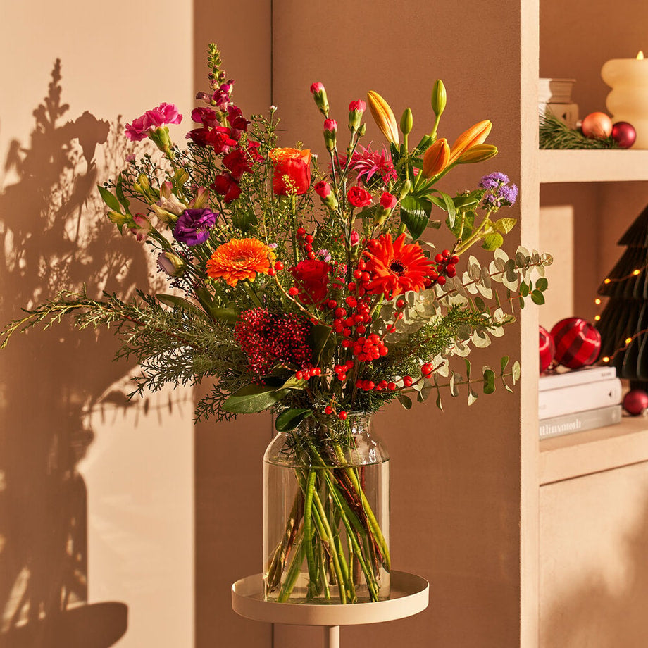 Kleurrijk kerstboeket met rode gerbera’s, oranje lelies en dennengroen in glazen vaas, met kerst details.
