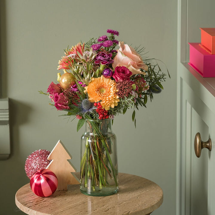 Kerstboeket met kleurrijke winterbloemen in glazen vaas in een feestelijke setting.