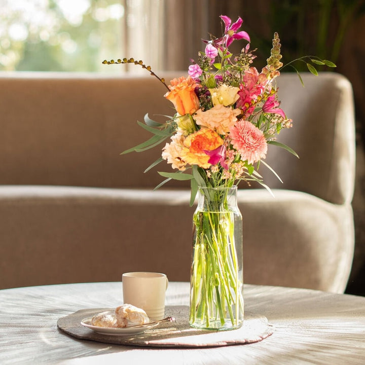 Kleurrijk boeketje met rozen en gerbera’s in een glazen vaasje op een tafel met koffie en koekjes.
