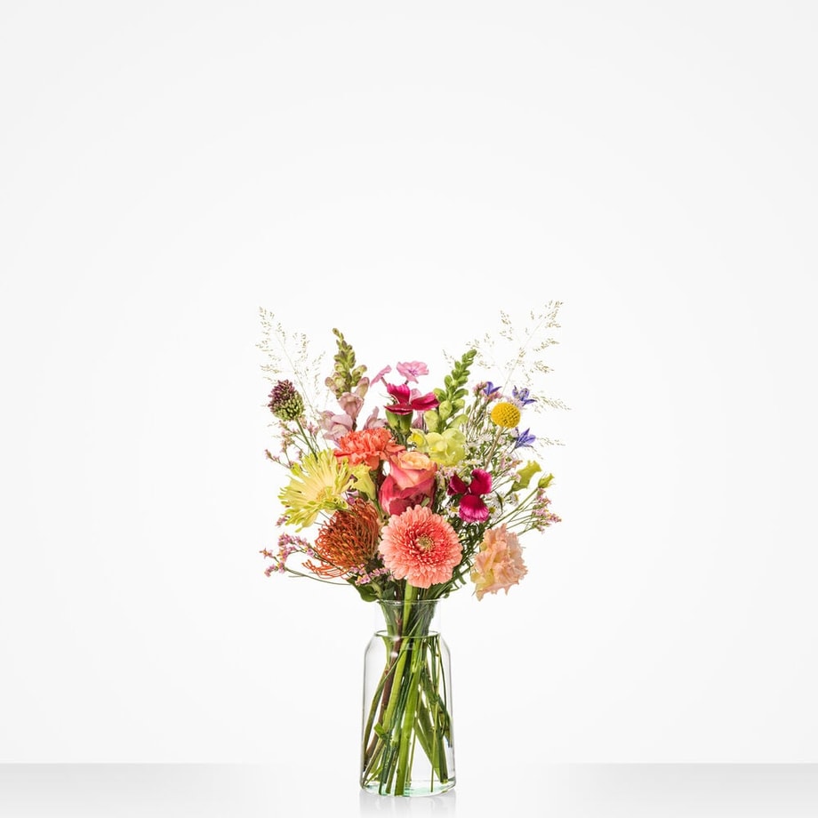Groot boeket vol kleurrijke bloemen zoals oranje gerbera’s, roze en rode rozen, in een hoge glazen vaas.