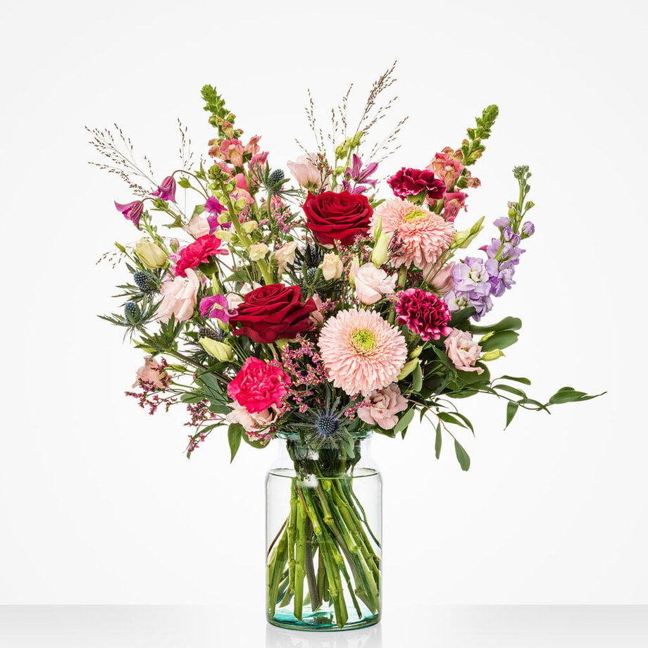 Een groot kleurrijk boeket met rode rozen, paarse bloemen, lichtroze dahlia’s en groen blad in een glazen vaas.