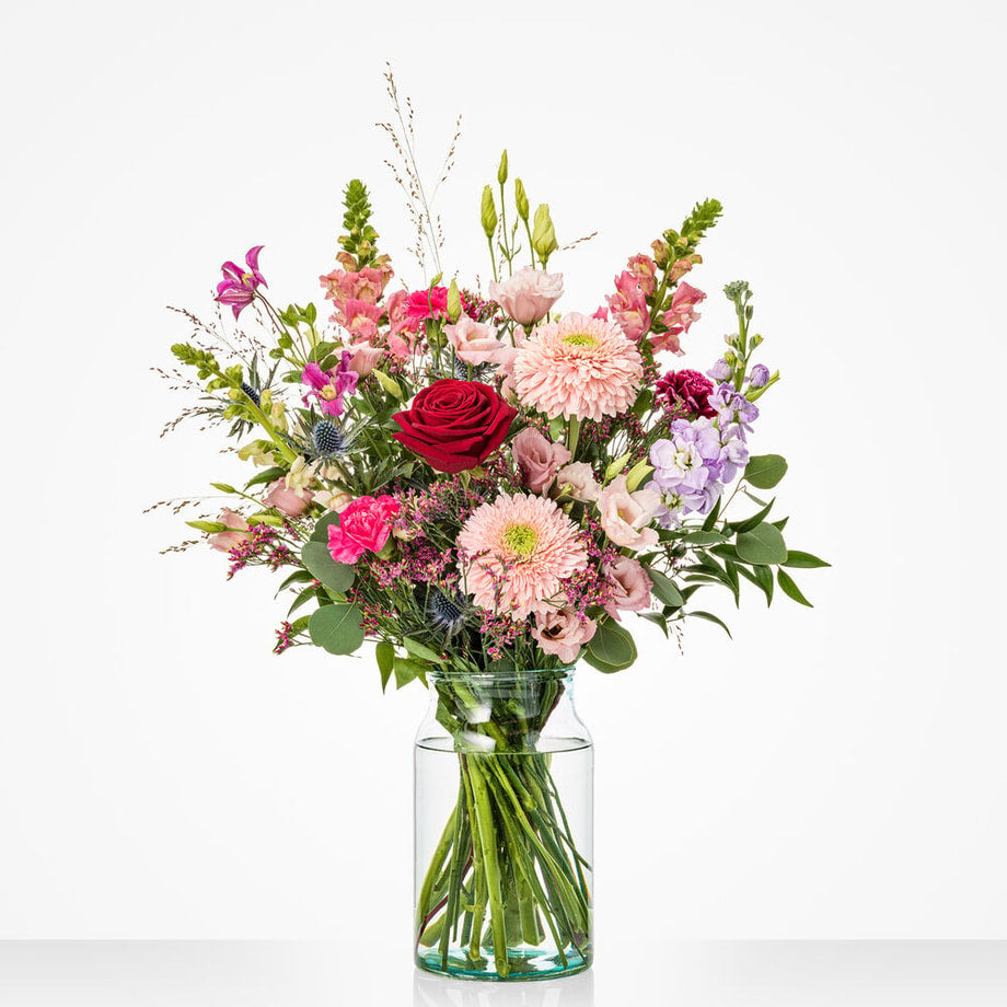 Een middelgroot boeket met roze dahlia’s, rode rozen en diverse andere bloemen in pasteltinten in een cilindervormige vaas.