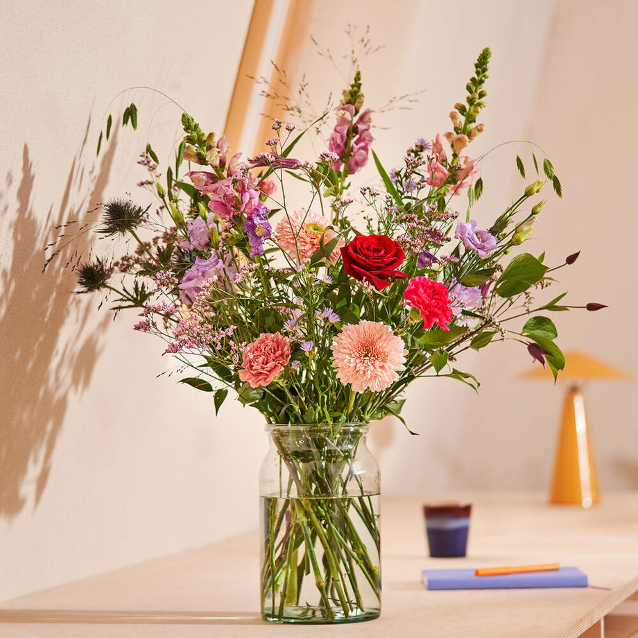 Een elegant boeket met een mix van rode, roze en paarse bloemen in een natuurlijke setting op een lichtgekleurde tafel in een minimalistische ruimte.