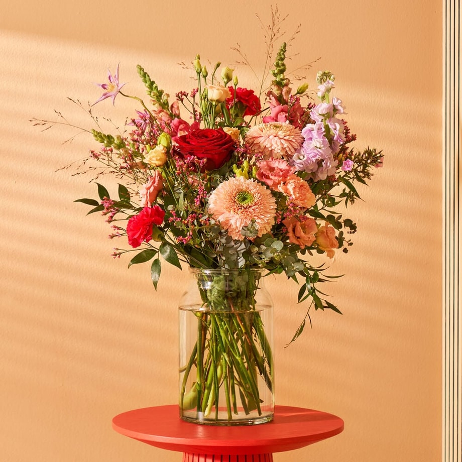 Een arrangement van roze, rode en lila bloemen, gemengd met groen, op een opvallend rood bijzettafeltje in een sfeervolle ruimte met warme tinten.