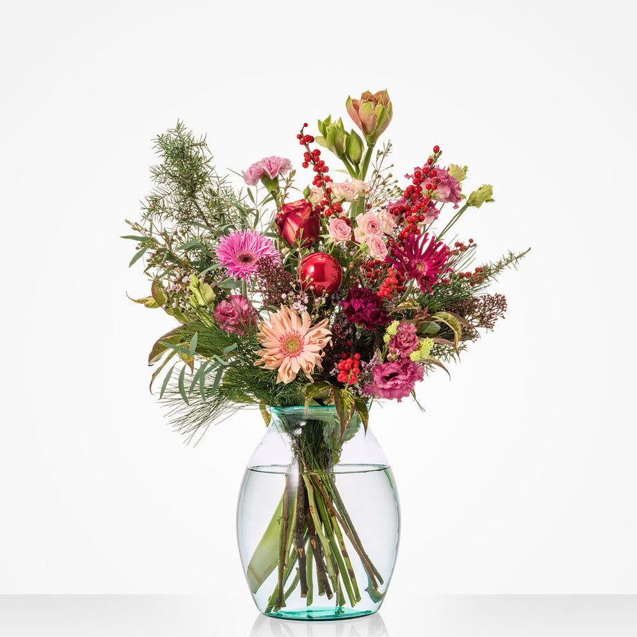 Klein kerstboeket met rozen, gerbera’s en rode kerstballen in glazen vaas.