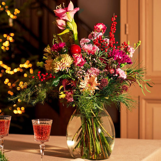 Kerstboeket met amaryllis, rozen en kerstdecoratie in een glazen vaas op een sfeervolle kersttafel.