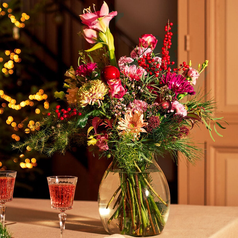 Kerstboeket met amaryllis, rozen en kerstdecoratie in een glazen vaas op een sfeervolle kersttafel.