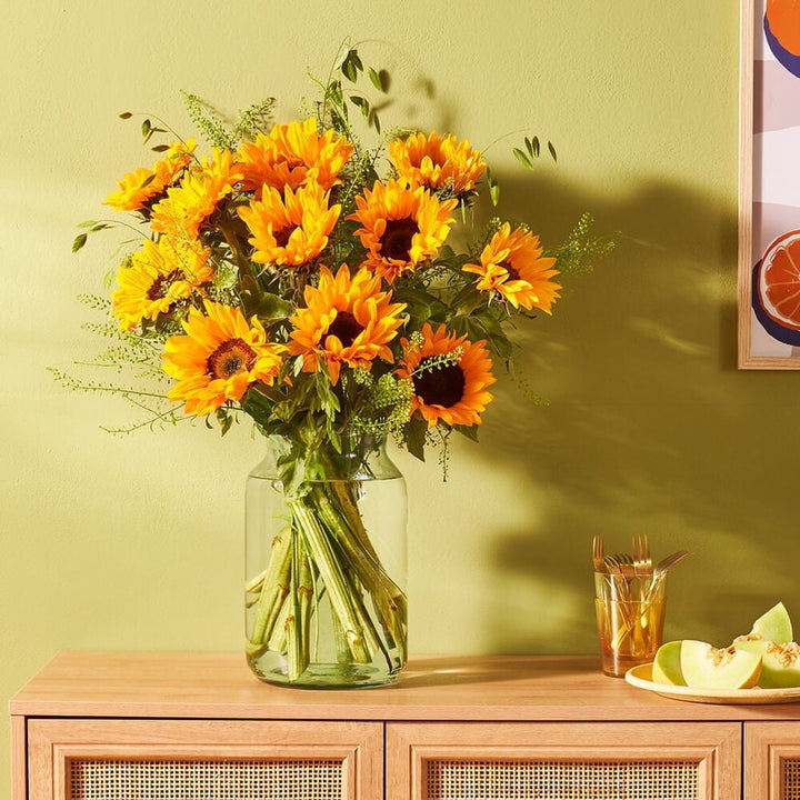 Groot boeket van zonnebloemen in een glazen vaas, geplaatst op een houten kast tegen een zachtgroene muur.