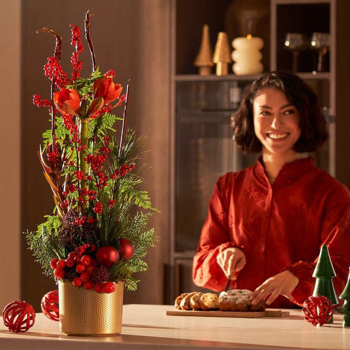 Luxe kerststuk met rode bessen, amaryllis en dennengroen in gouden pot, sfeervol geplaatst op een keukenblad naast een vrouw die kerststol snijdt.