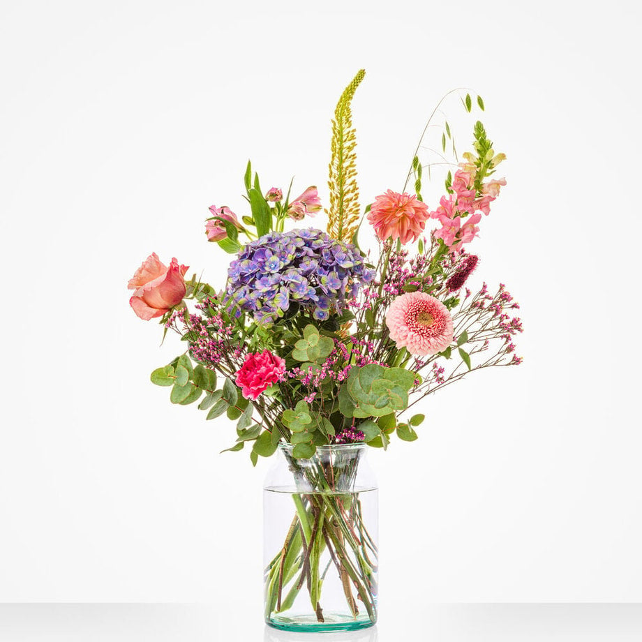 Klein maandboeket augustus met kleurrijke bloemen zoals roze gerbera’s, blauwe hortensia’s en lelies in een glazen vaas.
